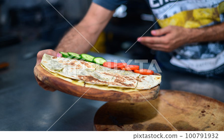 A man presents a wooden plate with a tortilla, cucumber, and tomatoes in the kitchen. 100791932