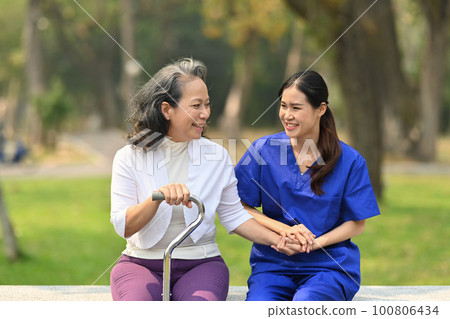 Smiling Asian female nurse holding senior woman hand having pleasant conversation during sitting outdoor in rehabilitation center Smiling Asian female nurse holding senior woman hand having pleasant conversation during sitting outdoor in rehabilitation center 100806434