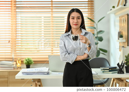 Young asian woman entrepreneur standing near working desk in a modern office, looking at the camera and smiling 100806472