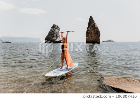 Woman sea sup. Close up portrait of happy young caucasian woman with long hair looking at camera and smiling. Cute woman portrait in bikini posing on sup board in the sea Woman sea sup. Close up portrait of happy young caucasian woman with long hair looking at camera and smiling. Cute woman portrait in bikini posing on sup board in the sea 100807410