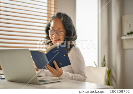 Relaxed and happy 60s Asian-aged woman reading a book at her workspace. 100809702