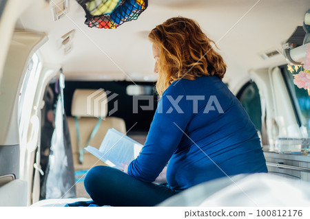 Side view middle-aged woman relaxing, reading book while sitting in camper car vehicle during road trip. Enjoying free lifestyle, travel, vacation, freedom. Selective focus 100812176