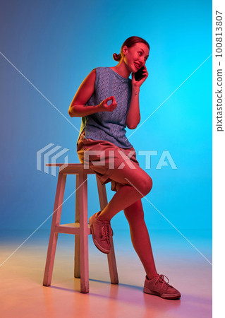 Portrait of young girl sitting on chair and talking via cell phone and telling joke over blue-pink background in neon light 100813807