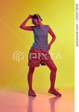 One young pretty girl wearing headphones standing, listening music and looking away over yellow-orange background in neon light One young pretty girl wearing headphones standing, listening music and looking away over yellow-orange background in neon light 100813808