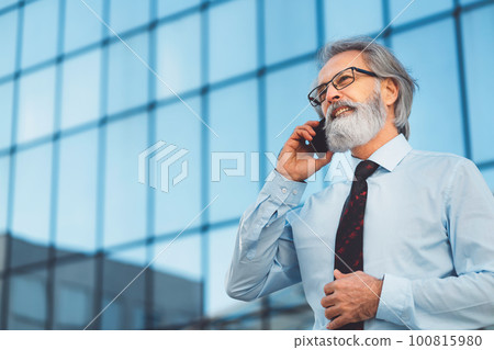 Low angle portrait of senior businessman standing outside with office building in the background, talking on the phone 100815980