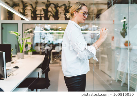 Side view of young blonde businesswoman with glasses writing on a clear board with white pen, working in modern glass office space Side view of young blonde businesswoman with glasses writing on a clear board with white pen, working in modern glass office space 100816030