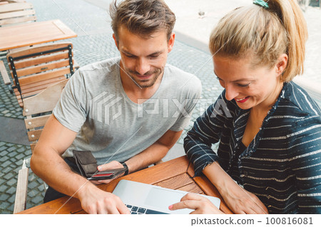 Young couple sitting at a bar on a sunny day with a laptop, working on something Young couple sitting at a bar on a sunny day with a laptop, working on something 100816081