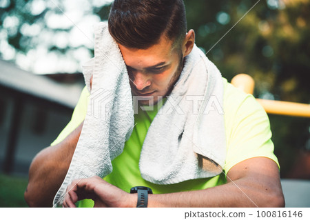 Sweaty fit man wiping him self with a towel after a workout while looking at his watch  100816146