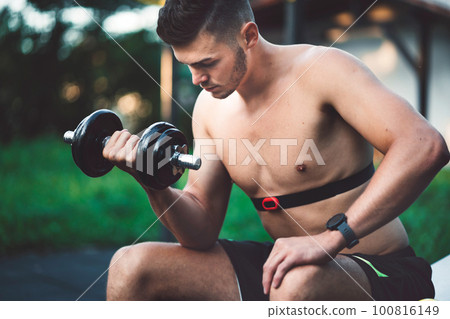 Fit caucasian man lifting weights outside, wearing a band around his chest to measure his pulse on the smart watch 100816149