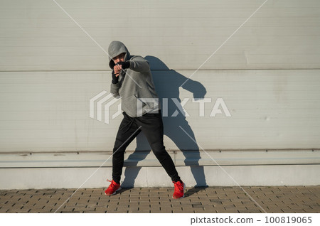 A man in a hoody trains boxing against a gray wall. A man in a hoody trains boxing against a gray wall. 100819065