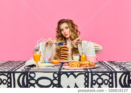 One charming woman gracefully holding cutlery and tasting pancakes over pink background. Pop art photography One charming woman gracefully holding cutlery and tasting pancakes over pink background. Pop art photography 100819137