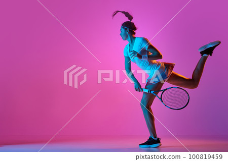 Serving ball with racket. Portrait of young woman, professional female tennis player in motion, training against pink studio background in neon light 100819459