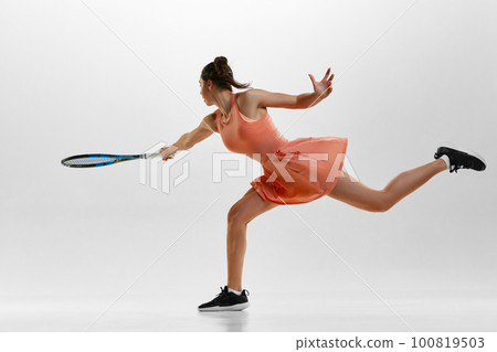 Serving ball, start of game. Portrait of young woman, professional female tennis player in uniform training against white studio background. 100819503