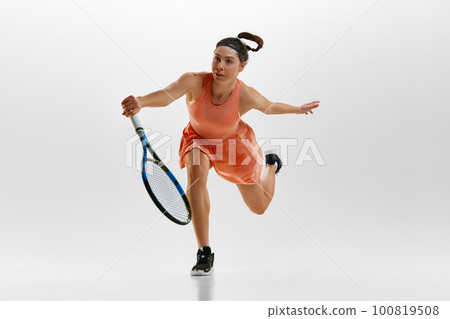 Portrait of young woman in motion, tennis player training against white studio background. Hitting ball with racket 100819508