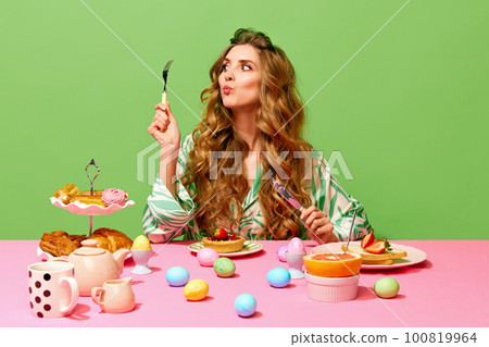 Food pop art of beautiful girl with long hair sitting at table holding cutlery and eating over green background. Colorful eggs on table Food pop art of beautiful girl with long hair sitting at table holding cutlery and eating over green background. Colorful eggs on table 100819964