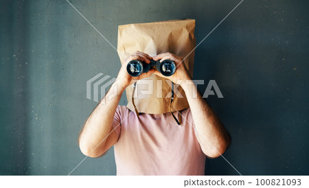 Man with a paper bag on head looking through binoculars to camera 100821093