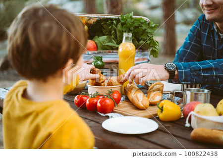 Father dad school kid boy child having a picnic in the forest camping site with vegetables, juice, coffee, and croissants. Wooden crate with fresh organic veggies surrounded with bread baguettes. 100822438