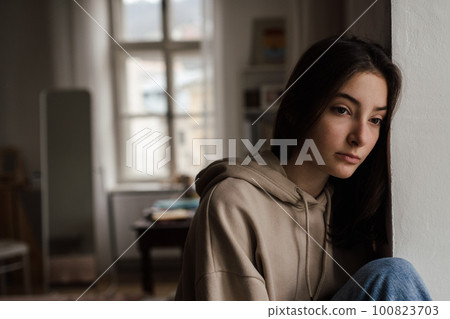 Unhappy teenage girl sitting in home and looking out of window. 100823703