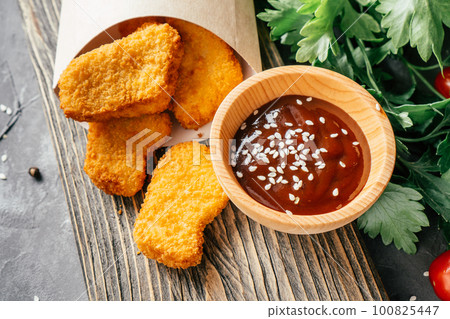 Serving of chicken nuggets in craft paper bag with barbecue sauce, sesame, cherry tomatoes, parsley on wooden surface Serving of chicken nuggets in craft paper bag with barbecue sauce, sesame, cherry tomatoes, parsley on wooden surface 100825447