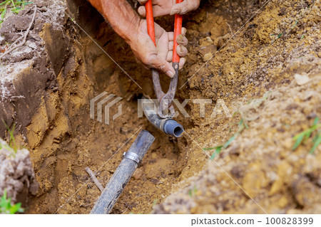 Man is working with pipes in ground while installing an underground sprinkler system to water garden on yard 100828399