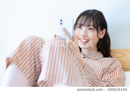 穿著睡衣的年輕女子在床上使用智能手機 穿著睡衣的年輕女子在床上使用智能手機 100829149