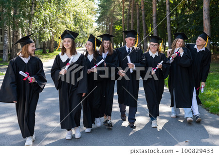 A group of graduates in robes with diplomas in their hands walk outdoors. Elderly student. 100829433