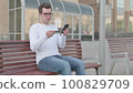 Young Man Reacting to Online Payment Failure on Smartphone while Sitting Outdoor on Bench 100829709