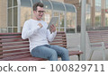 Young Man Celebrating Online Win on Tablet while Sitting Outdoor on Bench 100829711