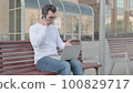 Young Man Talking on Phone and using Laptop while Sitting Outdoor on Bench 100829717
