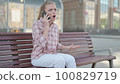 Angry Young Woman Talking on Phone while Sitting Outdoor on Bench 100829719
