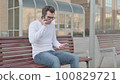 Angry Young Man Talking on Phone while Sitting Outdoor on Bench 100829721