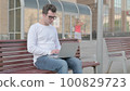 Busy Young Man Using Laptop Sitting Outdoor on Bench 100829723