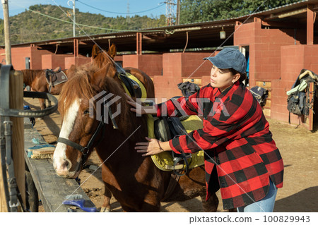Portrait of positive young female worker in plaid shirt standing close to saddled horse in countryside club 100829943