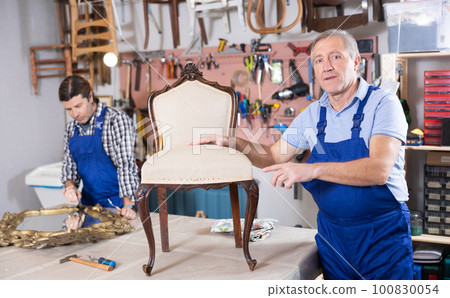Experienced male restorer showing restored vintage chair in workshop 100830054