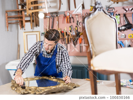 collected male restorer carefully covers carved wooden frame of antique mirror with layer of varnish 100830166