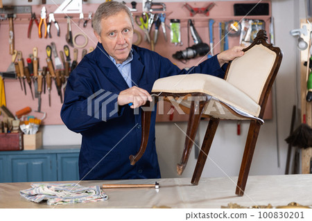 Elderly male furniture maker working on vintage chair in workshop Elderly male furniture maker working on vintage chair in workshop 100830201