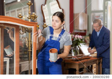 Young female furniture workshop worker repainting vintage glass cabinet Young female furniture workshop worker repainting vintage glass cabinet 100830280