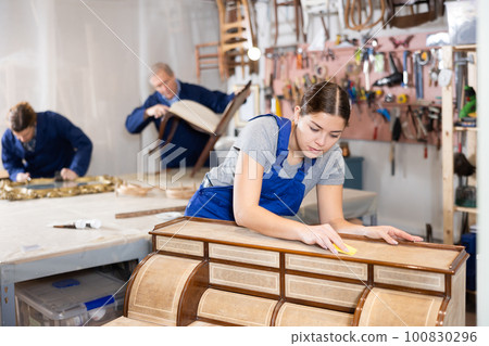 Woman restorer carefully and carefully restores chest of drawers in restoration workshop 100830296