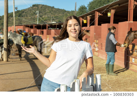Portrait of young female stable visitor smiling at camera in horse club on sunny spring day 100830303