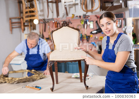 Skilled female furniture repair specialist in uniform restoring Victorian chair during workday in workshop 100830363