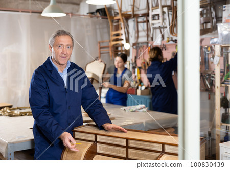 Ederly craftsman polishes tabletop of old chest of drawers with rounded drawers with soft cloth 100830439