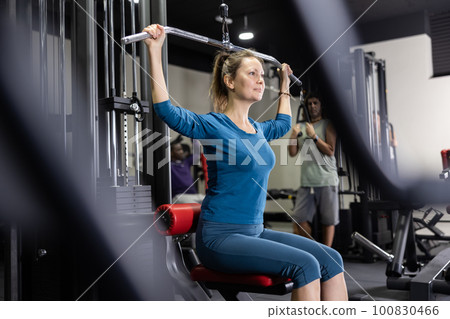 Caucasian woman doing exercises on lat pull down machine in gym Caucasian woman doing exercises on lat pull down machine in gym 100830466