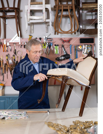 Professional elderly craftsman restorer in uniform renewing antique chair while working in repair shop Professional elderly craftsman restorer in uniform renewing antique chair while working in repair shop 100830500