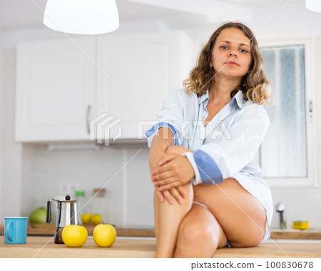 Lovely sexy girl in shirt posing while sitting on the table of modern kitchen 100830678