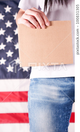 Young woman holds empty cardboard with Space for Text sign against American flag on background. Girl protesting anti-abortion laws. Feminist power. Womens rights freedom. Young woman holds empty cardboard with Space for Text sign against American flag on background. Girl protesting anti-abortion laws. Feminist power. Womens rights freedom. 100831305