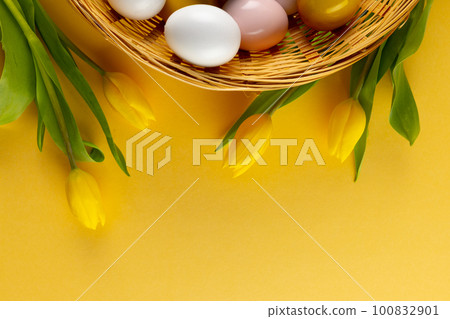 Image of multi coloured easter eggs in basket with yellow tulips and copy space on yellow background 100832901