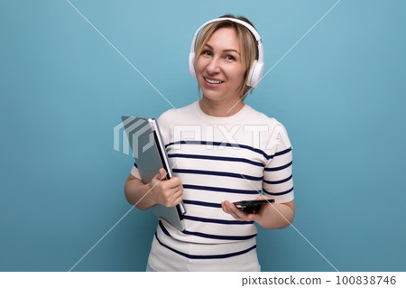 portrait of cute smiling blond young female student with big wireless headphones and laptop on blue portrait of cute smiling blond young female student with big wireless headphones and laptop on blue 100838746