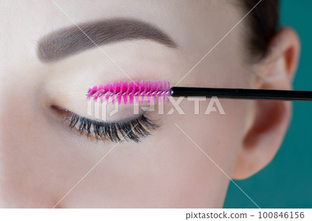 Closed eye with long eyelashes, black eyeliner, perfect skin eyebrow of unrecognizable young woman holding pink spoolie. Closed eye with long eyelashes, black eyeliner, perfect skin eyebrow of unrecognizable young woman holding pink spoolie. 100846156