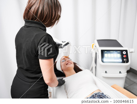 Cosmetologist using diode laser device while performing rejuvenation facial treatment. Woman lying near cosmetology equipment while having face lifting procedure. Focus on female doctor. 100846615