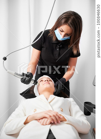 Female cosmetologist in medical mask using professional beauty device while performing face lifting procedure in cosmetology clinic. Woman receiving radiofrequency facial treatment in clinic. 100846630